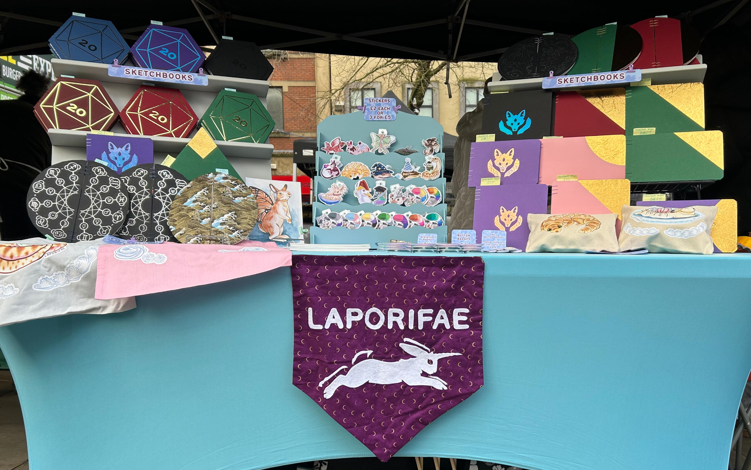 A table with sketchbooks, books, and stickers displayed. The table has a banner that says Laporifae.